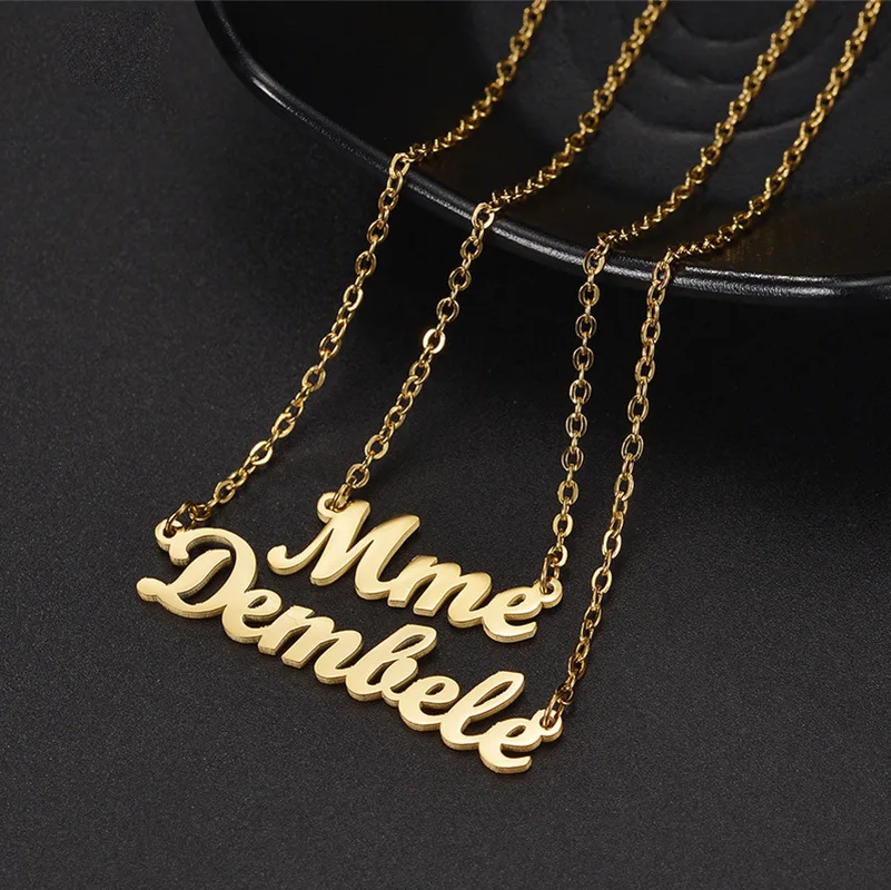 Gold necklace with "Mme Dembele" pendant on a dark background.