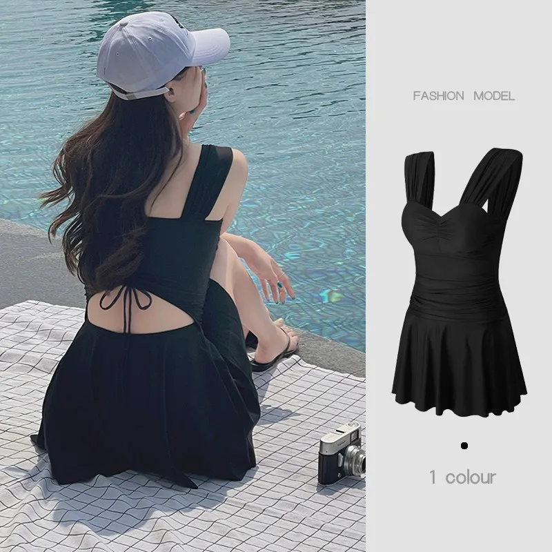 Woman in a stylish black dress by the water, wearing a white cap.