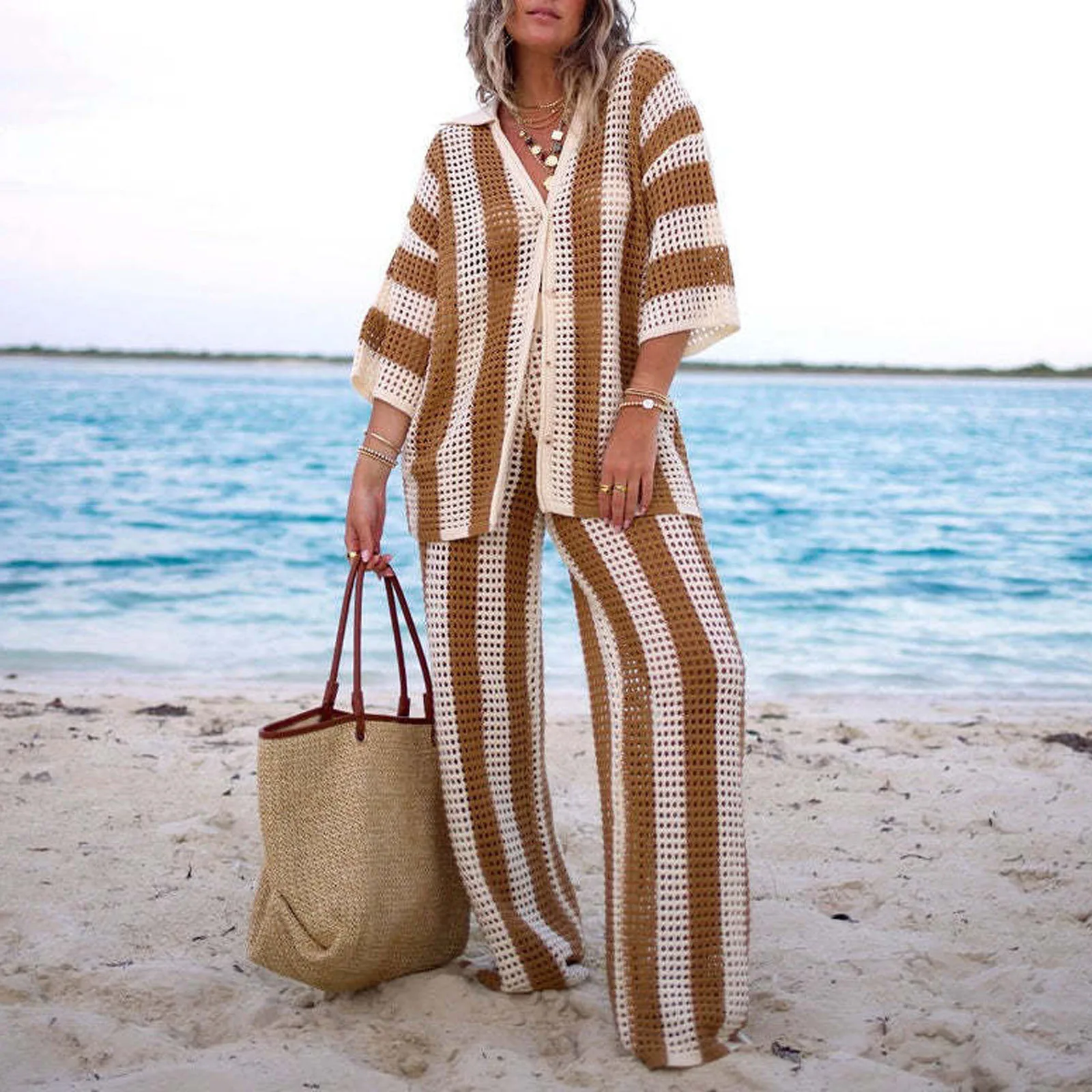 Woman in a striped beach outfit carrying matching bags by the ocean.
